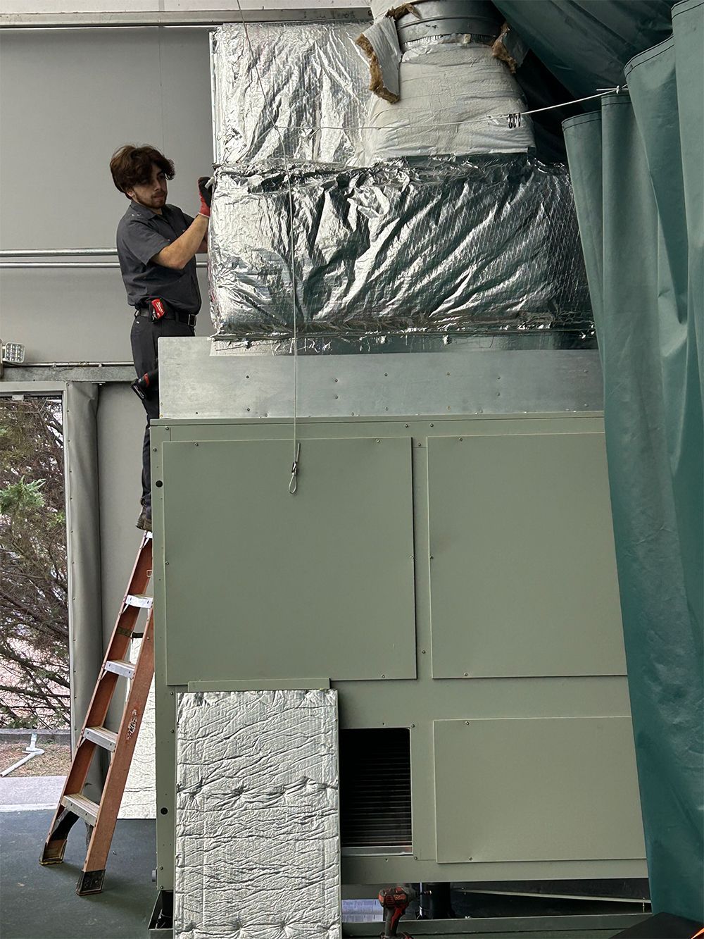 Person on a ladder working on HVAC unit, covered in foil insulation. Grey and silver. Indoors.
