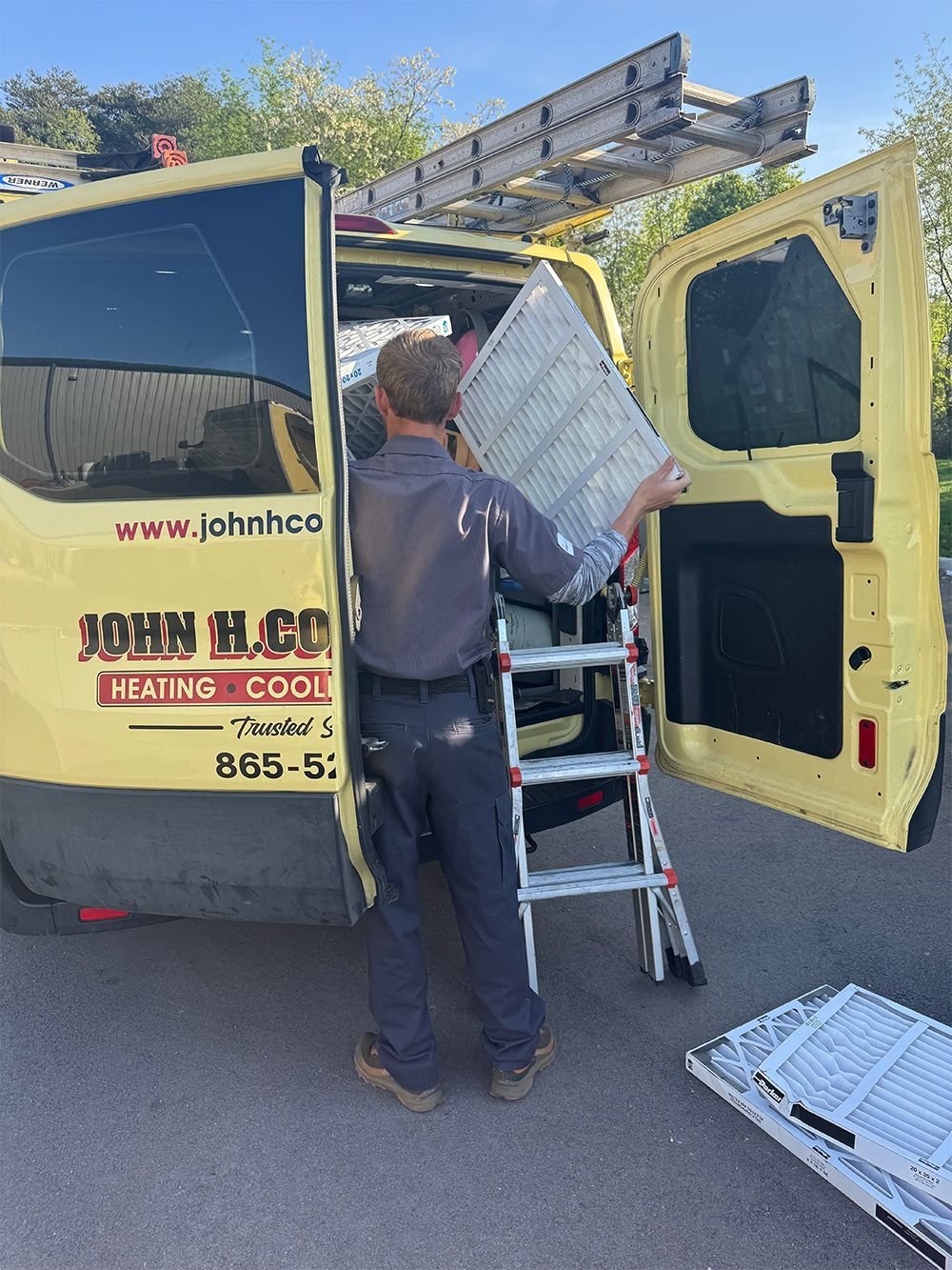 Man loading HVAC filter into yellow work van. Exterior, sunny day. Van door open, ladder beside.