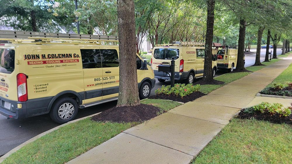 An electrician in a yellow hard hat is using a multimeter on electrical components in a grey box, outdoors.