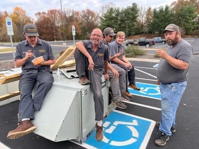 Five HVAC workers take a break near a handicap parking space. Two eat, one points, all sit on unit.