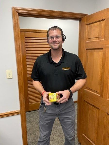 Man in black polo shirt and gray pants holds a small yellow box, standing in a doorway.