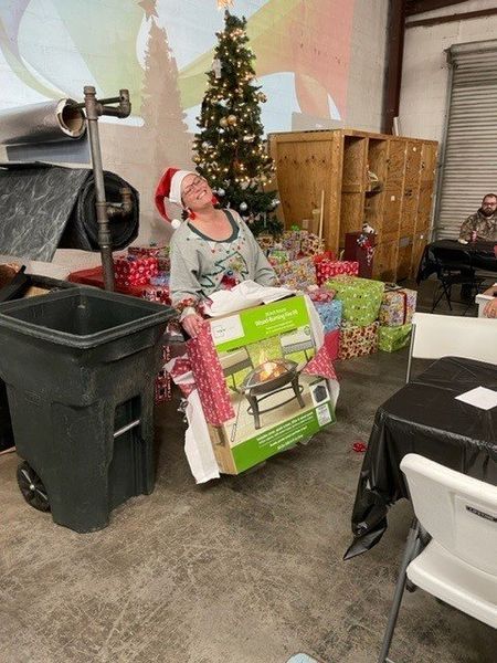 Person wearing Santa hat surrounded by gifts and a Christmas tree. In a room with trash can and furniture.