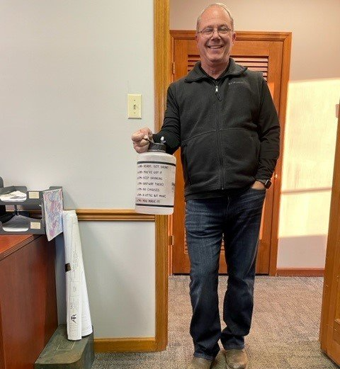 Man holding a jar with text in an office setting, smiling. He wears a black jacket and jeans.