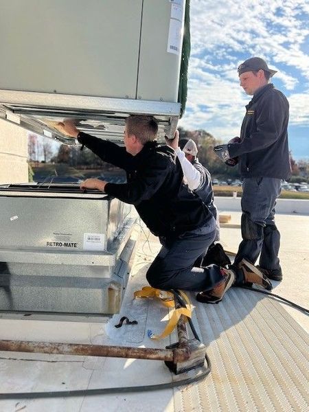 Two workers installing equipment on a rooftop. One kneels and works while the other watches.