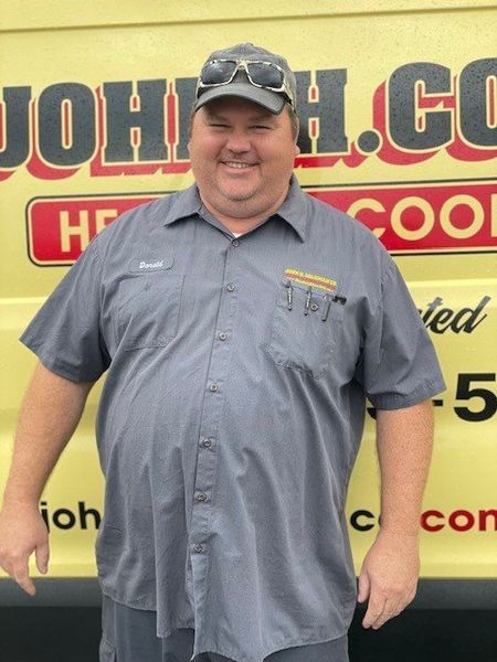 Man in gray work shirt, hat with sunglasses on top, smiling in front of a yellow van with company logo.