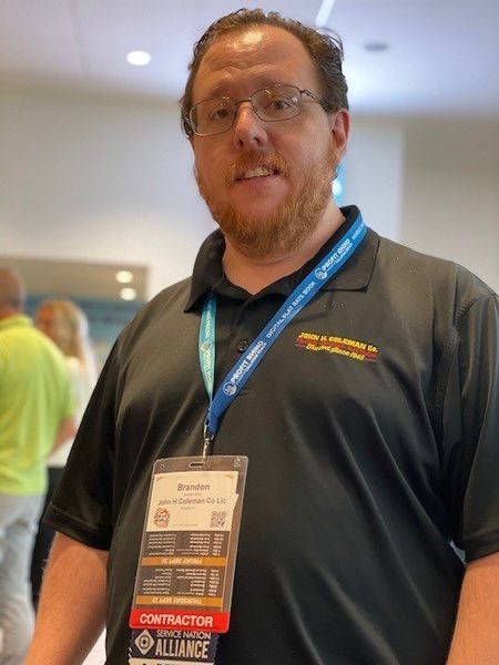 Man wearing glasses and a black polo shirt, with a name badge, standing indoors.