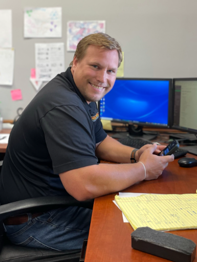 Man at desk smiling, holding a device. Blue computer monitor behind him.