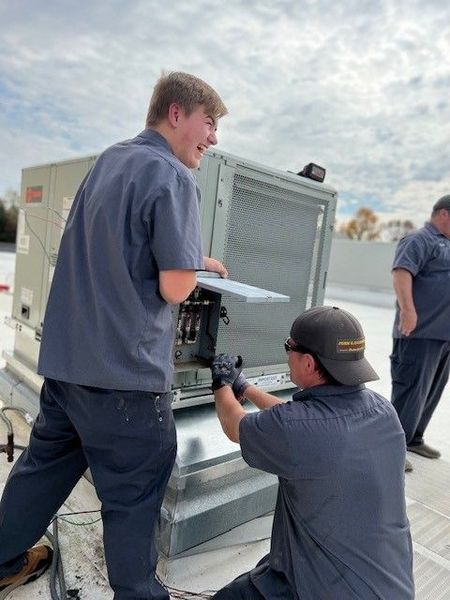 Three HVAC technicians working on a rooftop unit. One kneels, working inside, while the other two stand nearby.