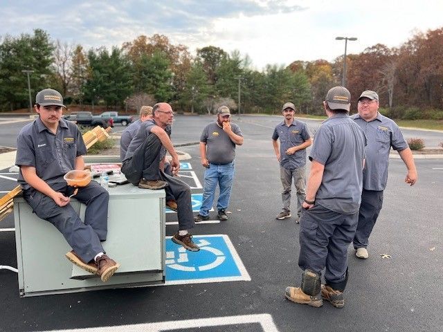 Group of workers in gray uniforms, gathered outdoors in parking lot, near equipment. Cloudy sky overhead.