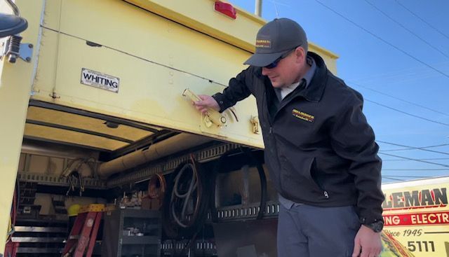 Man in black jacket opening compartment on a yellow service truck, outdoors.
