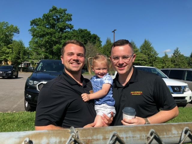 Two men and a toddler smiling outside; one man holds the child. Cars and trees in the background.