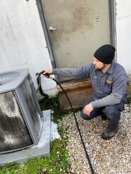Man cleaning an AC unit outside a building with a hose. Gray and black tones.
