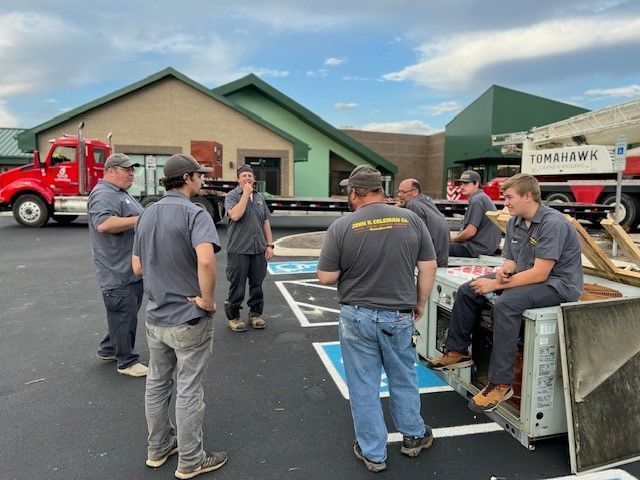 A group of workers in gray shirts and jeans are gathered outdoors near a building and parked trucks.