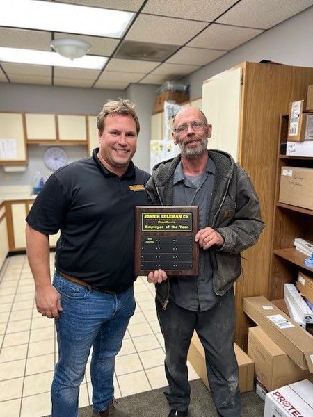Man presents award plaque to another man. They stand indoors by cabinets.