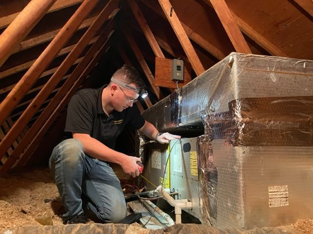 A person in an attic inspecting HVAC equipment. They wear safety glasses and a headlamp.
