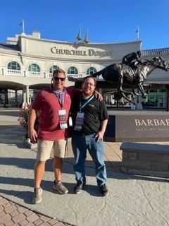 Two men pose in front of Churchill Downs; one in red shirt, khaki shorts, the other in black shirt, jeans.