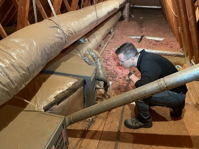 Man inspecting HVAC system in an attic. He's holding a flashlight, examining ducts and insulation.