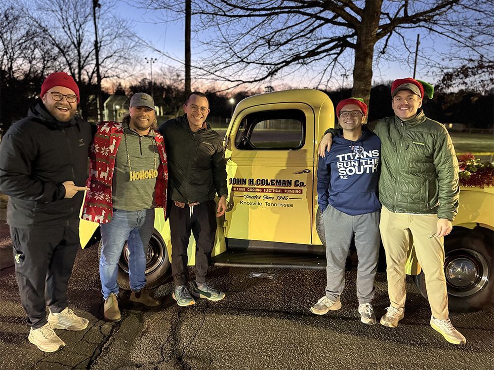 Five people stand in front of a yellow vintage truck; all wear red hats. Evening setting.