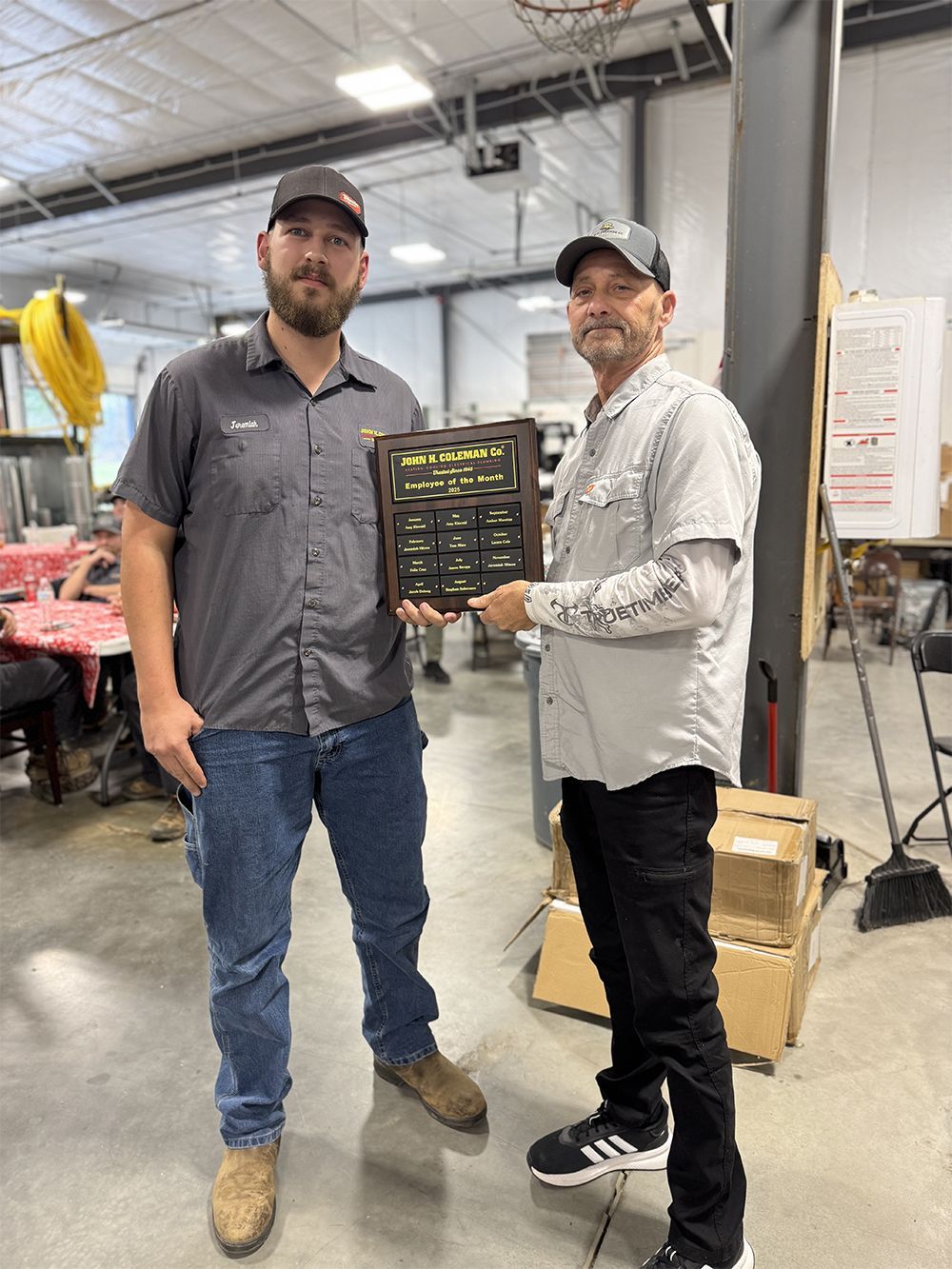 Two men in a shop, one holding a plaque. The man on the left is wearing a hat, shirt and jeans. The other man is wearing a hat, shirt and black pants.