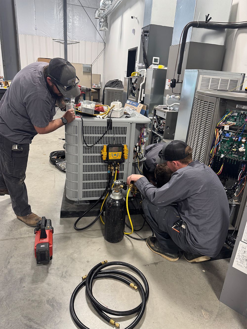 Two technicians work on an air conditioning unit indoors. One kneels, connecting a hose. The other leans over the unit.