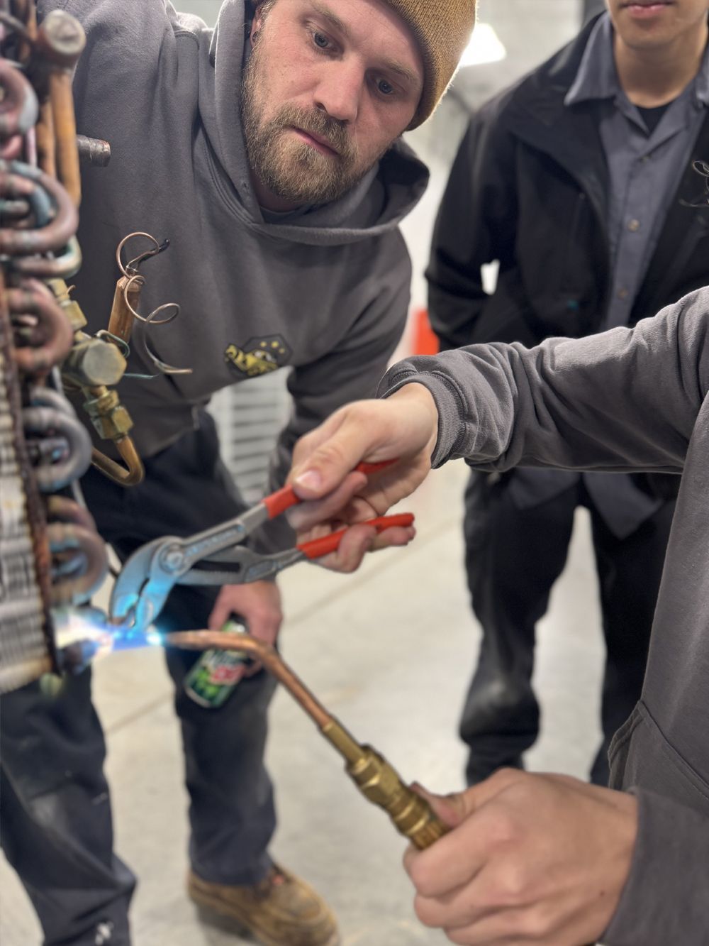 Man soldering copper pipes with focused expression, indoors. Another man watches.