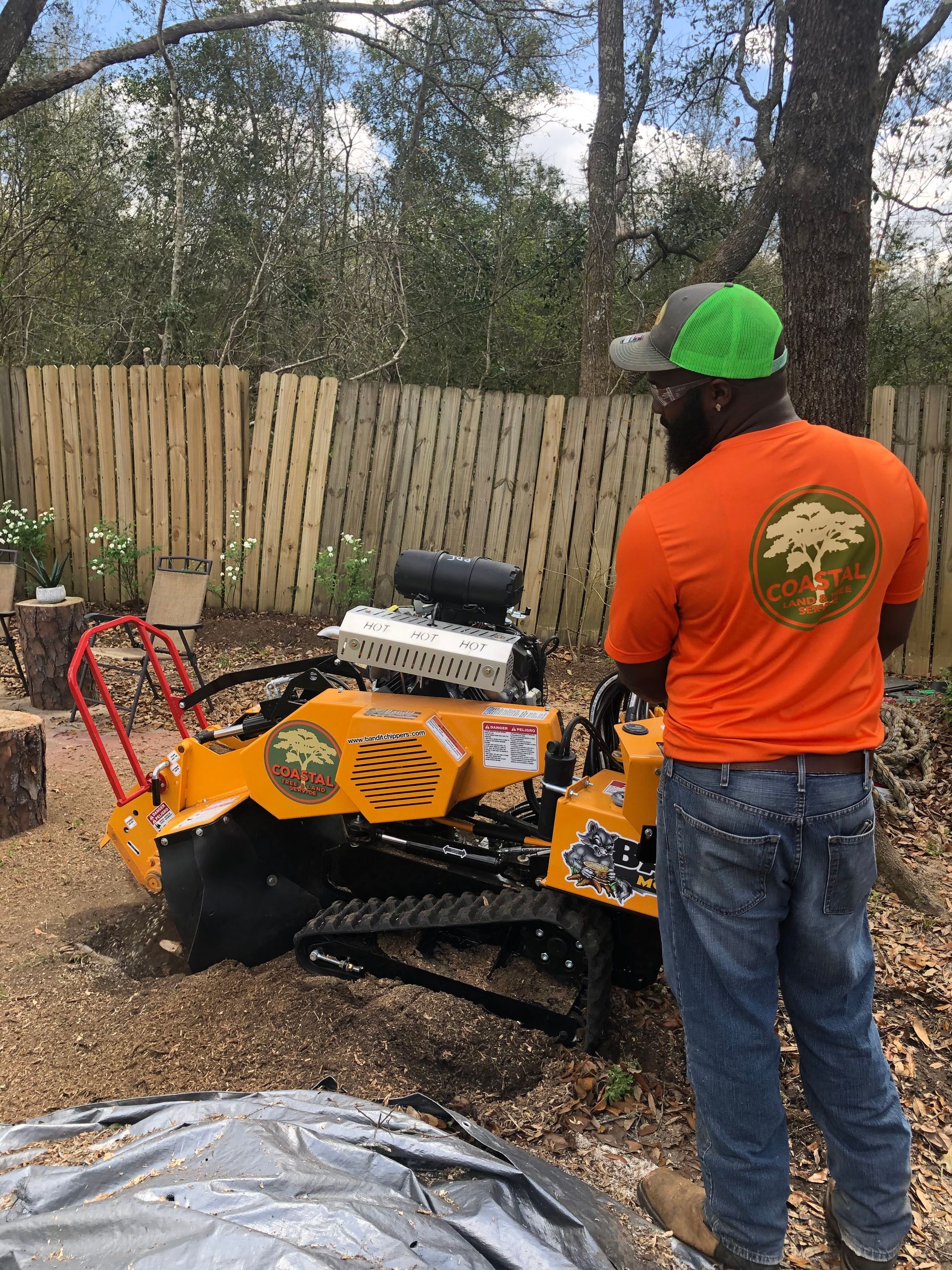 A man in an orange shirt is standing next to a tree stump grinder.