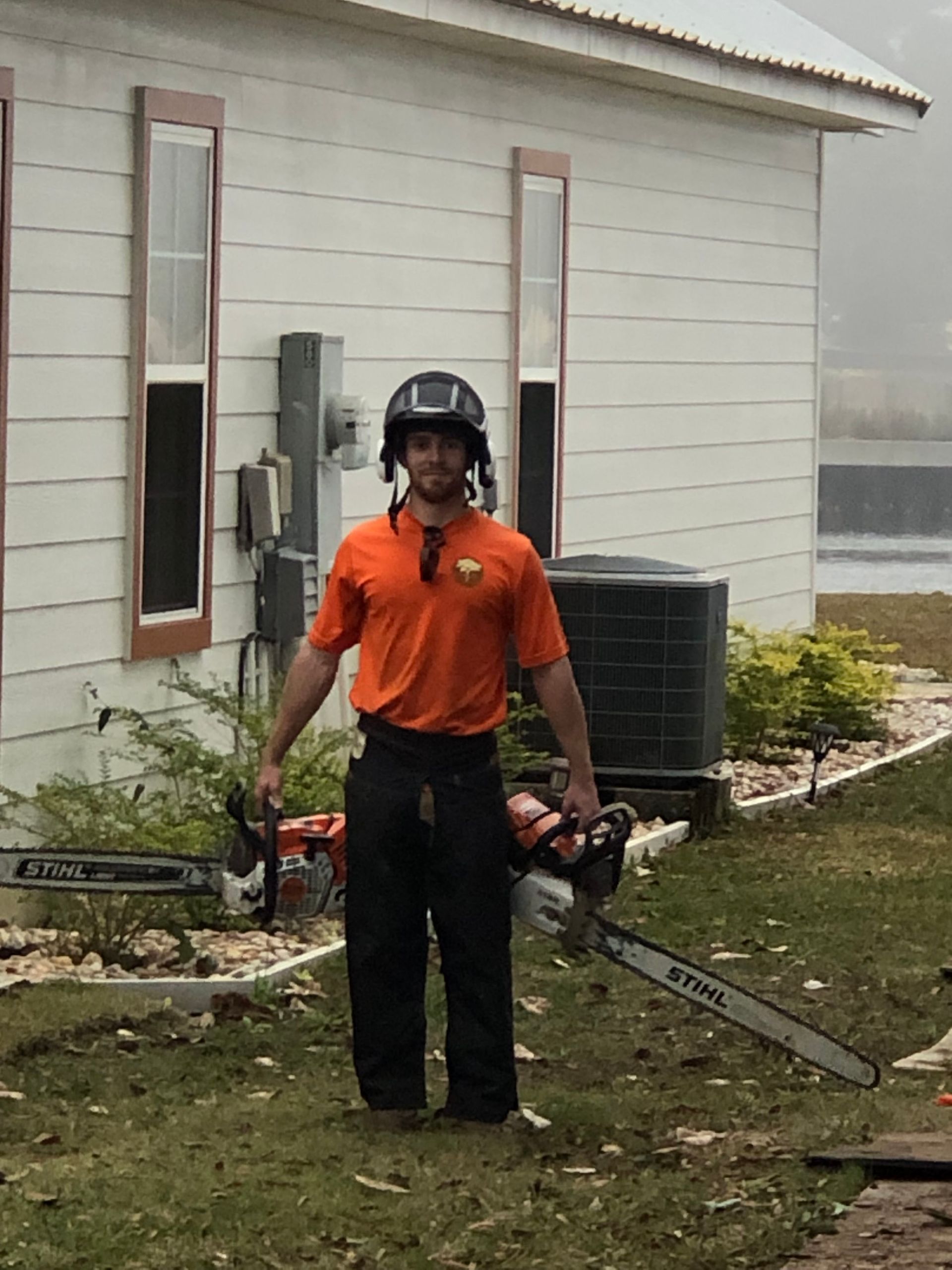 A man in an orange shirt is holding a chainsaw in front of a house