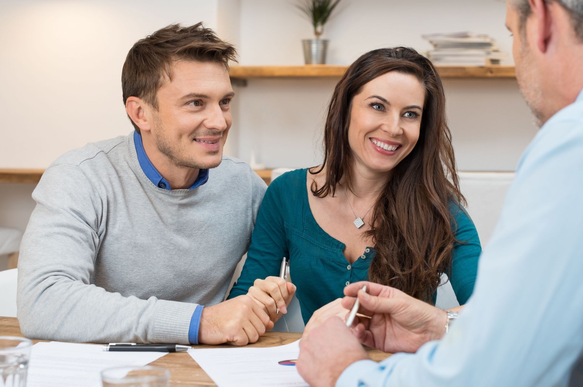 Couple smiles while holding hands, meeting with a person at a table, discussing documents.
