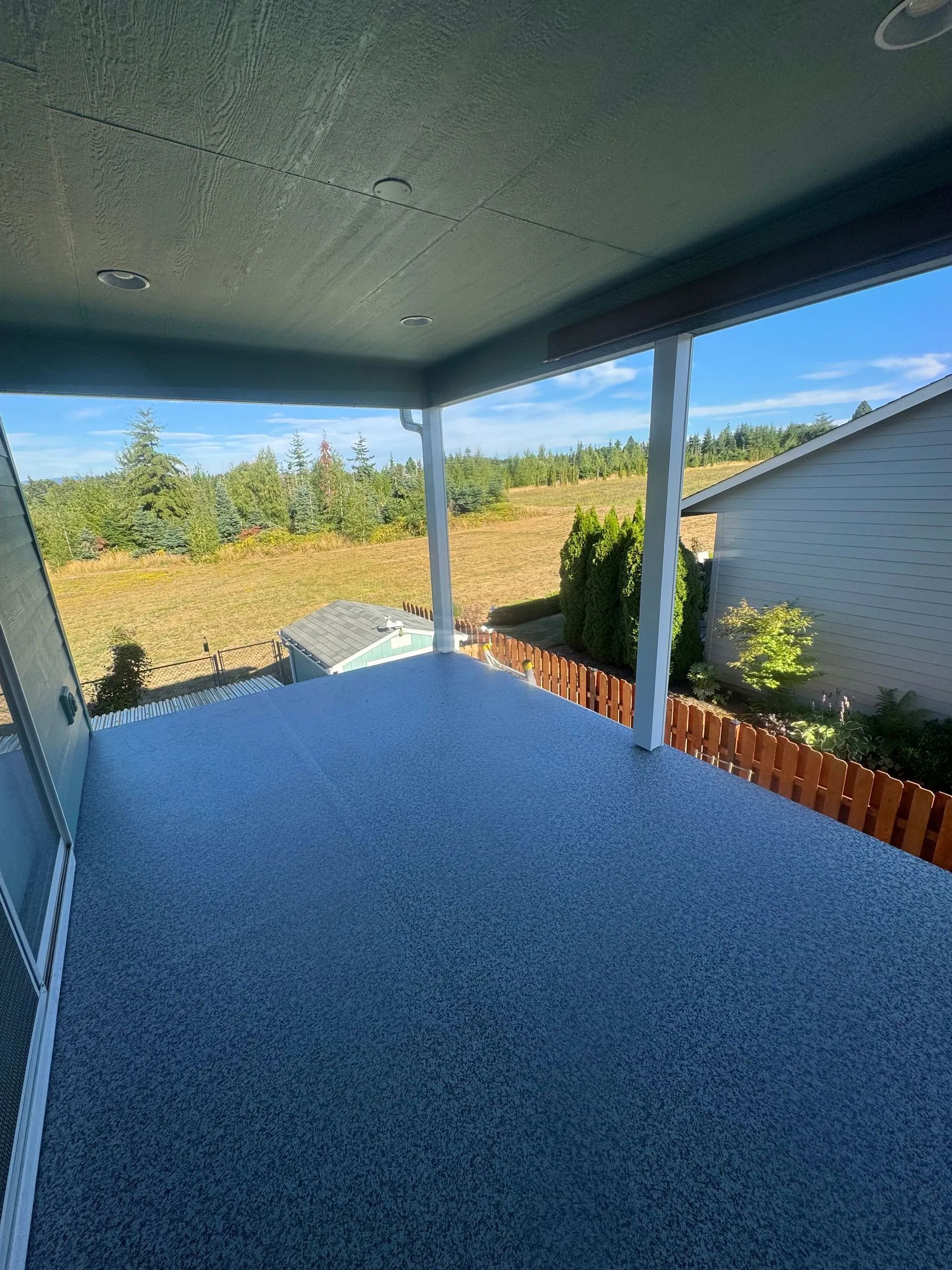 Covered patio with blue flooring, white posts, and a view of trees and a neighboring house beyond a brick fence.
