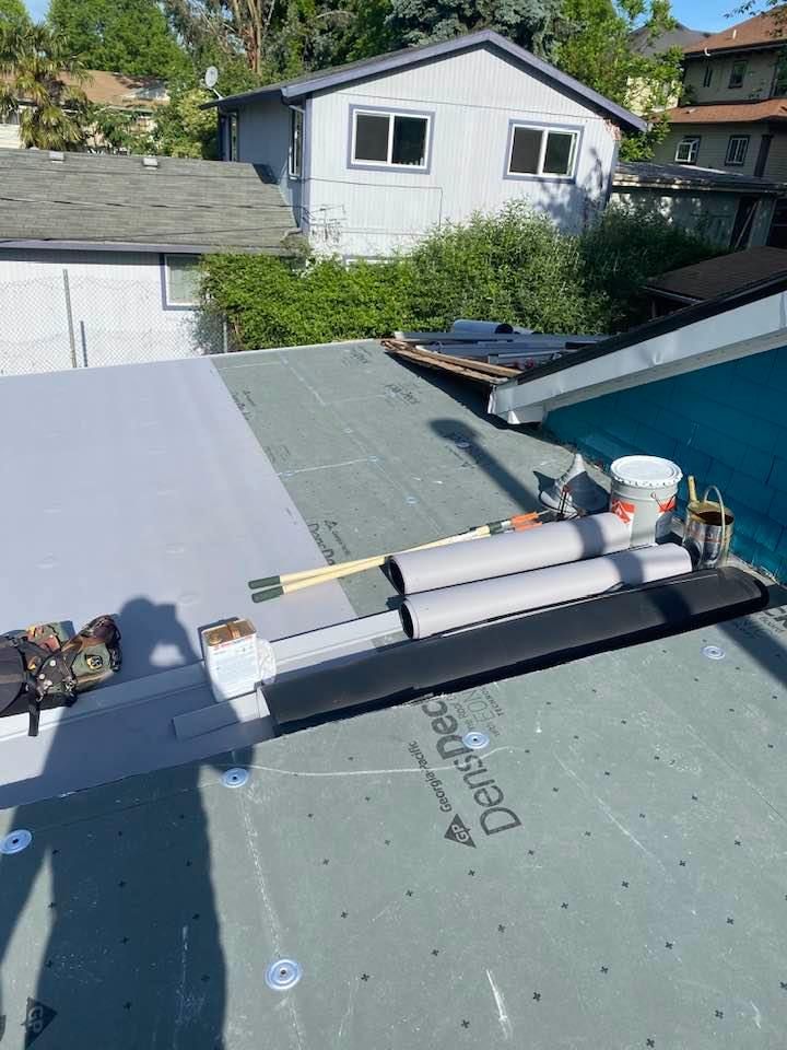 Rooftop under repair with rolled roofing, tools, and buckets on a flat roof near a house