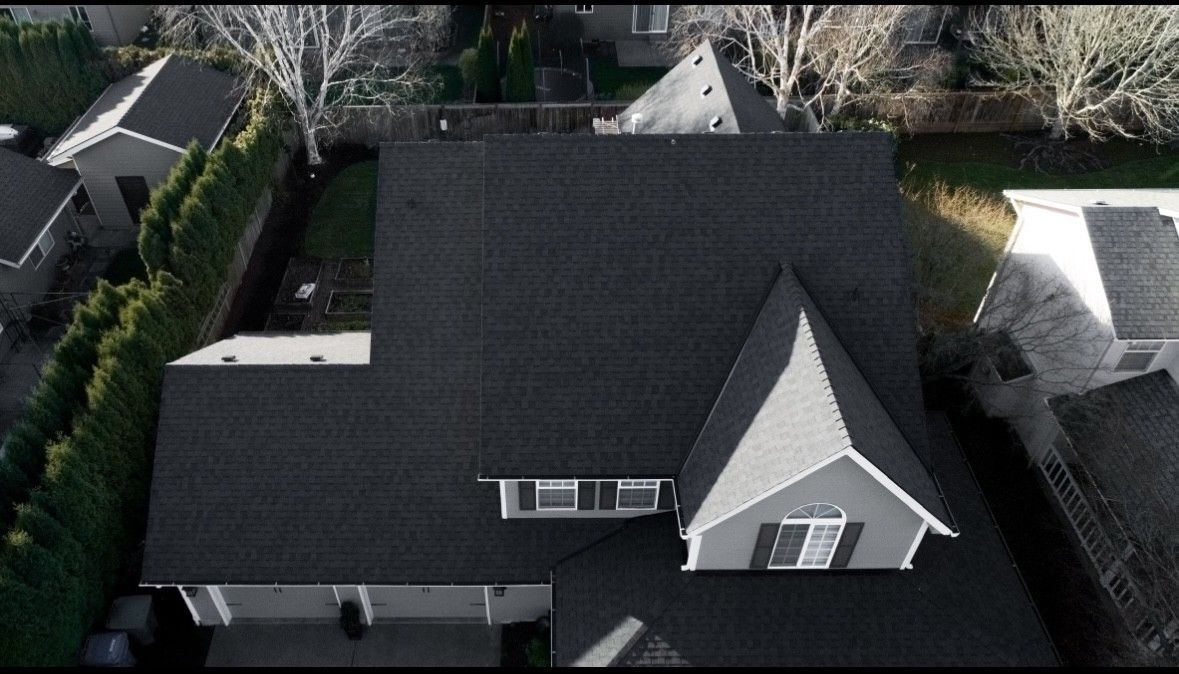 Aerial view of a dark shingled house roof with dormer and surrounding trees and neighboring homes.