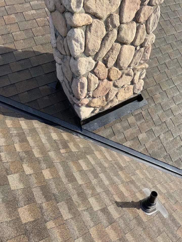 Stone chimney on a shingled roof with a vent pipe nearby