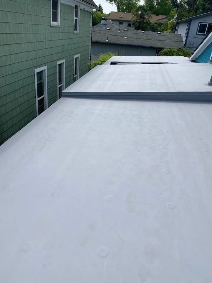 White flat roof beside a green-sided house, with a raised seam and neighboring rooftops in view