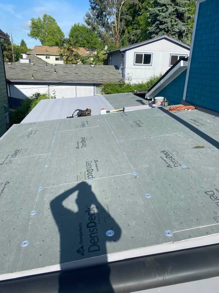 Flat roof under repair with gray roofing membrane, shadow of person, and houses and trees in the background