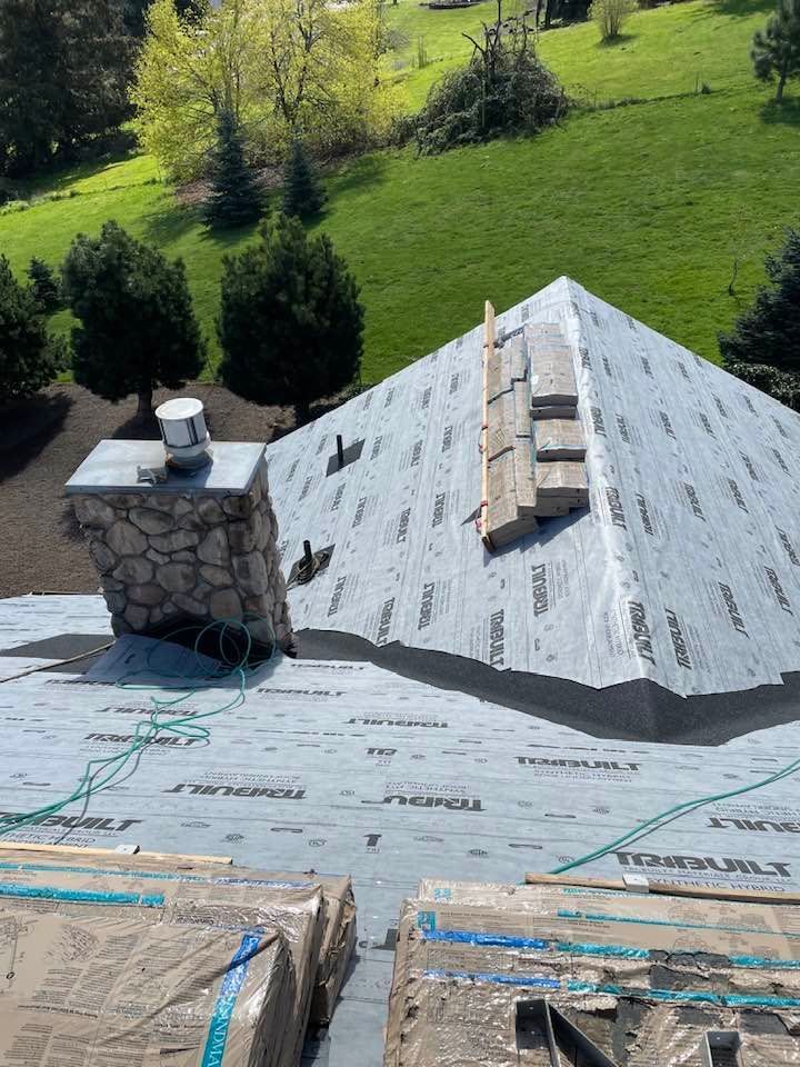 Roof under construction with gray shingles, vent pipe, and stone chimney beside a green lawn.