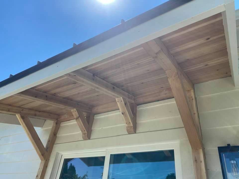 Wooden porch roof with exposed beams over a house window under a bright blue sky