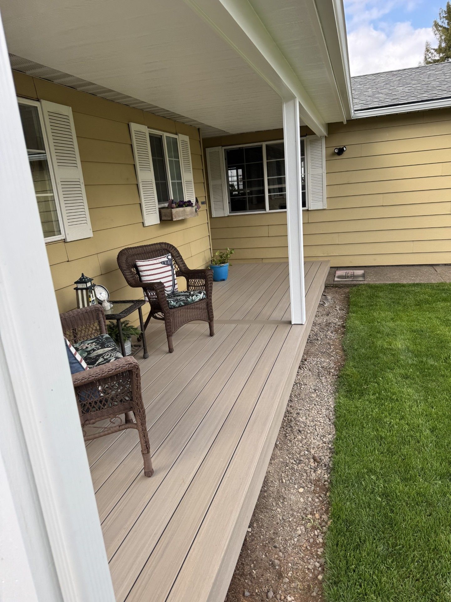 Covered front porch with beige siding, white posts, wicker chairs, and a potted plant beside a lawn.