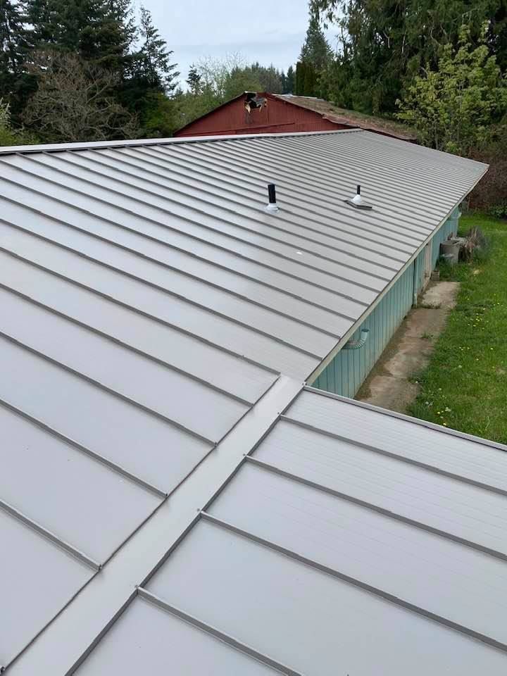Gray metal roof with standing seams and vent pipes, viewed from above near a grassy yard and trees
