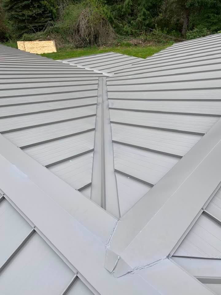 Gray metal roof with intersecting seams and a central valley, viewed from above amid green trees