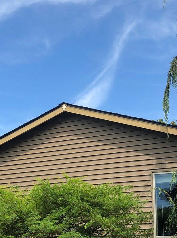 The top of a tan-sided house with a dark roof against a bright blue sky with a faint, wispy cloud.