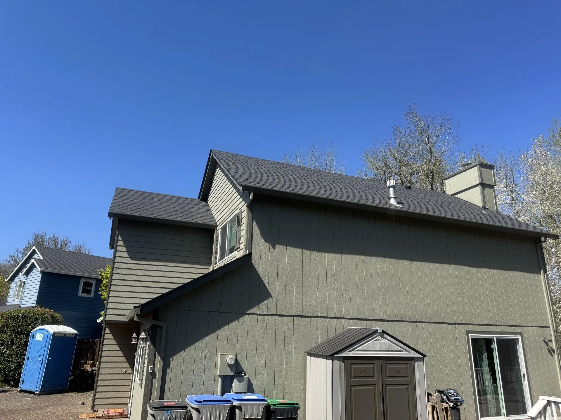 Gray house with a dark roof, small porch, and blue recycling bin under a clear blue sky