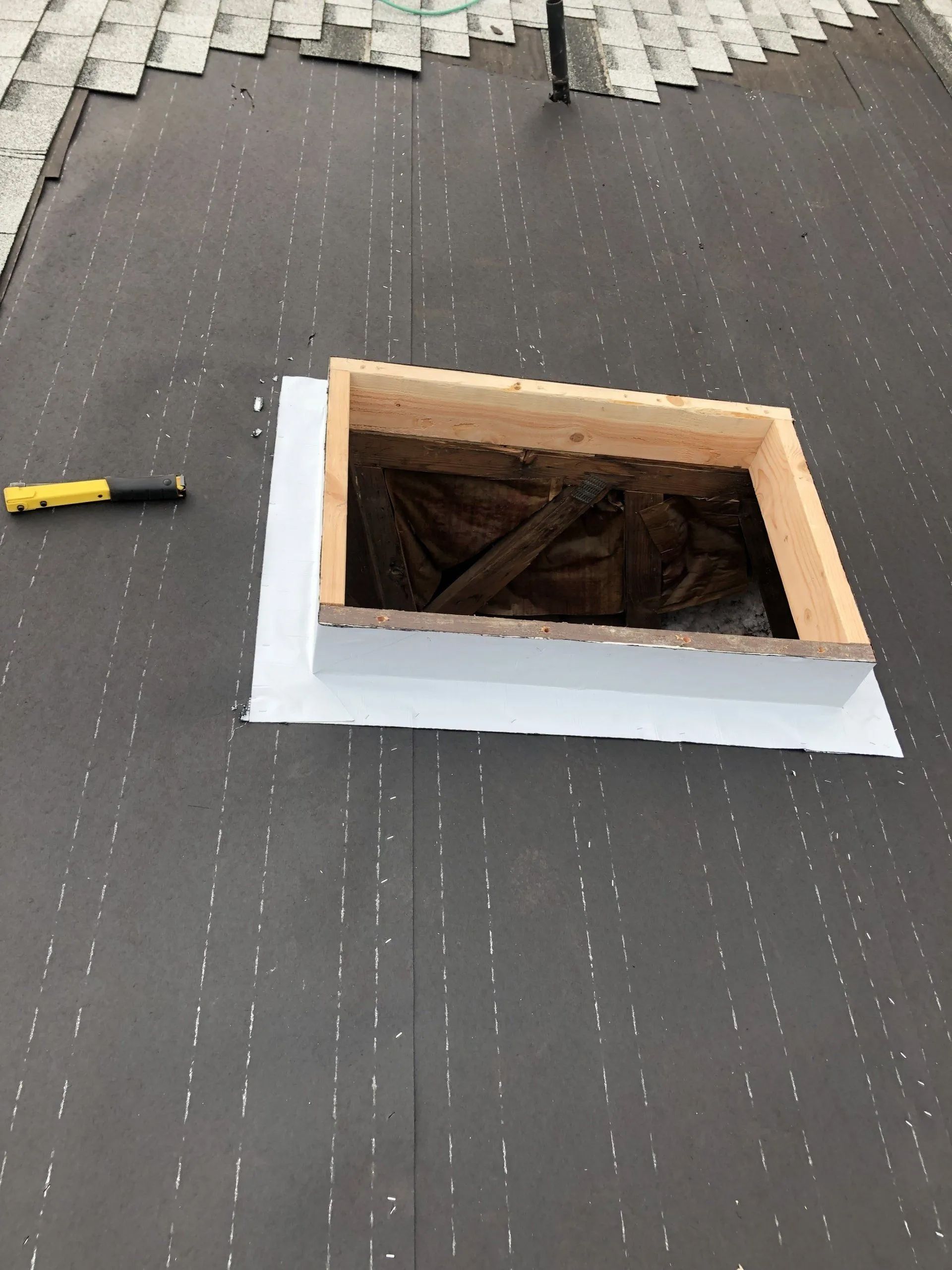 Flat roof with a square skylight opening, white flashing, and a yellow-handled tool nearby.