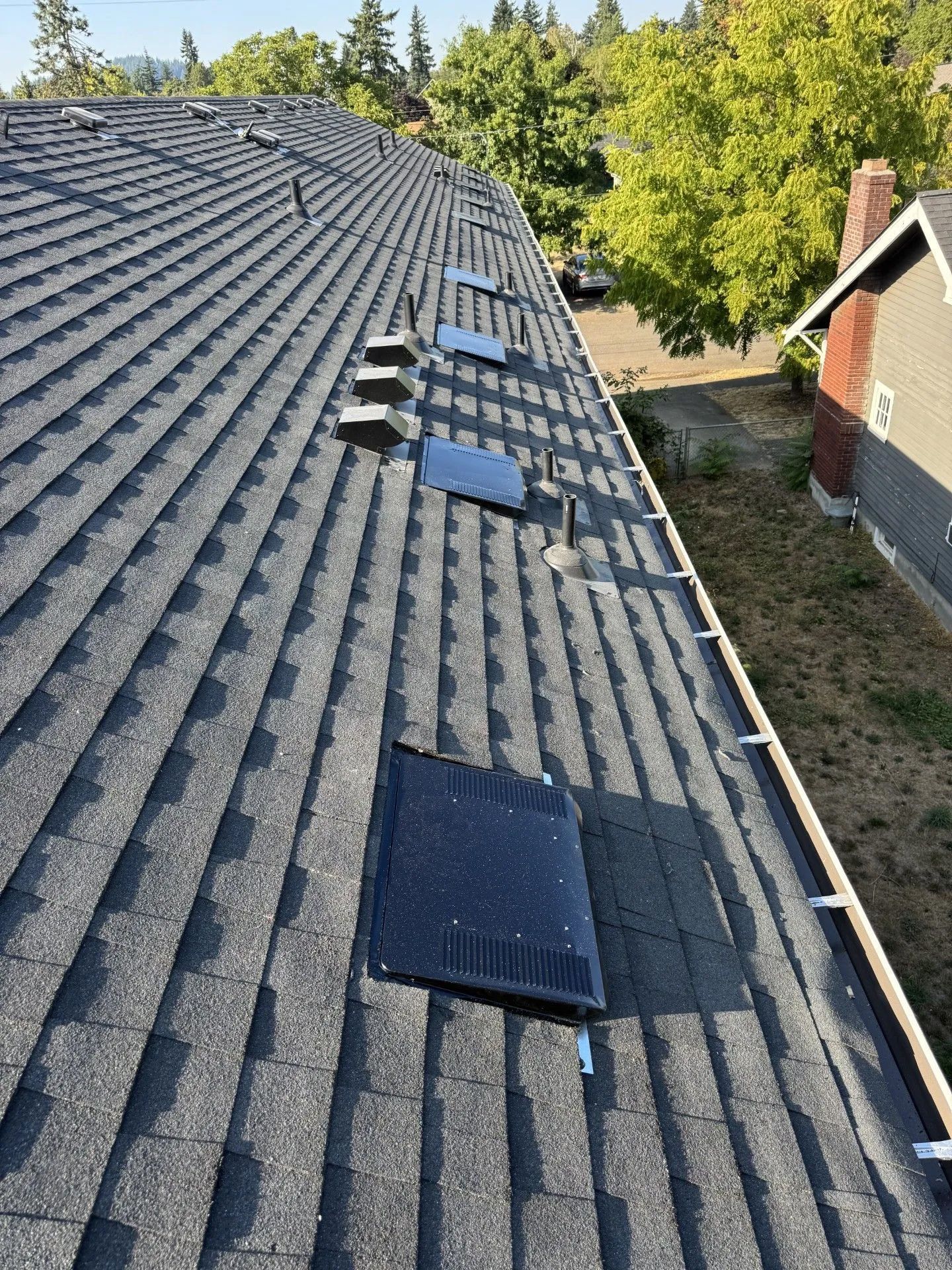 Roof with dark asphalt shingles and several solar panels, viewed from above near trees and a house.