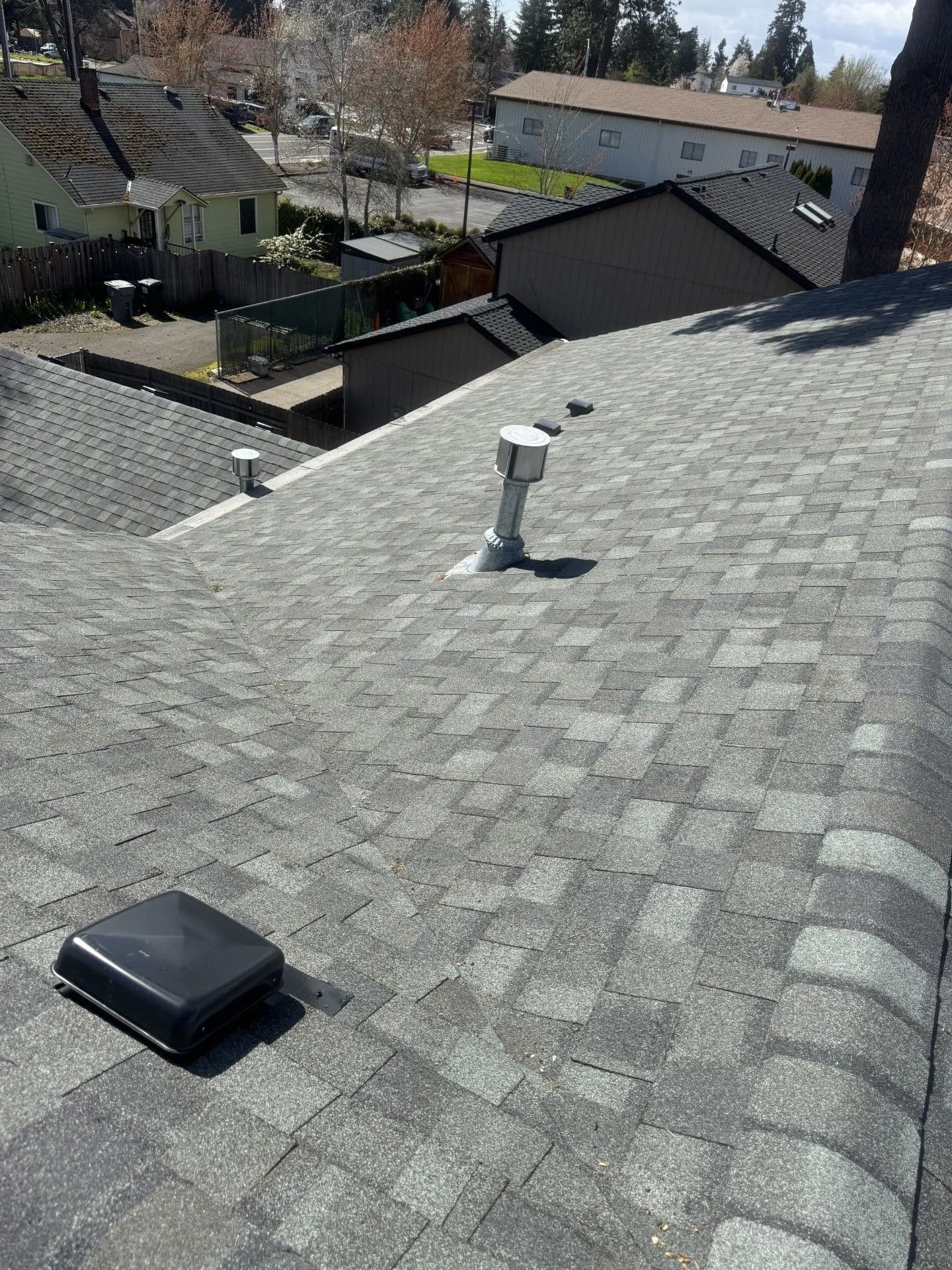 Rooftop with gray shingles, vent pipe, and black skylight, overlooking neighboring houses and trees.