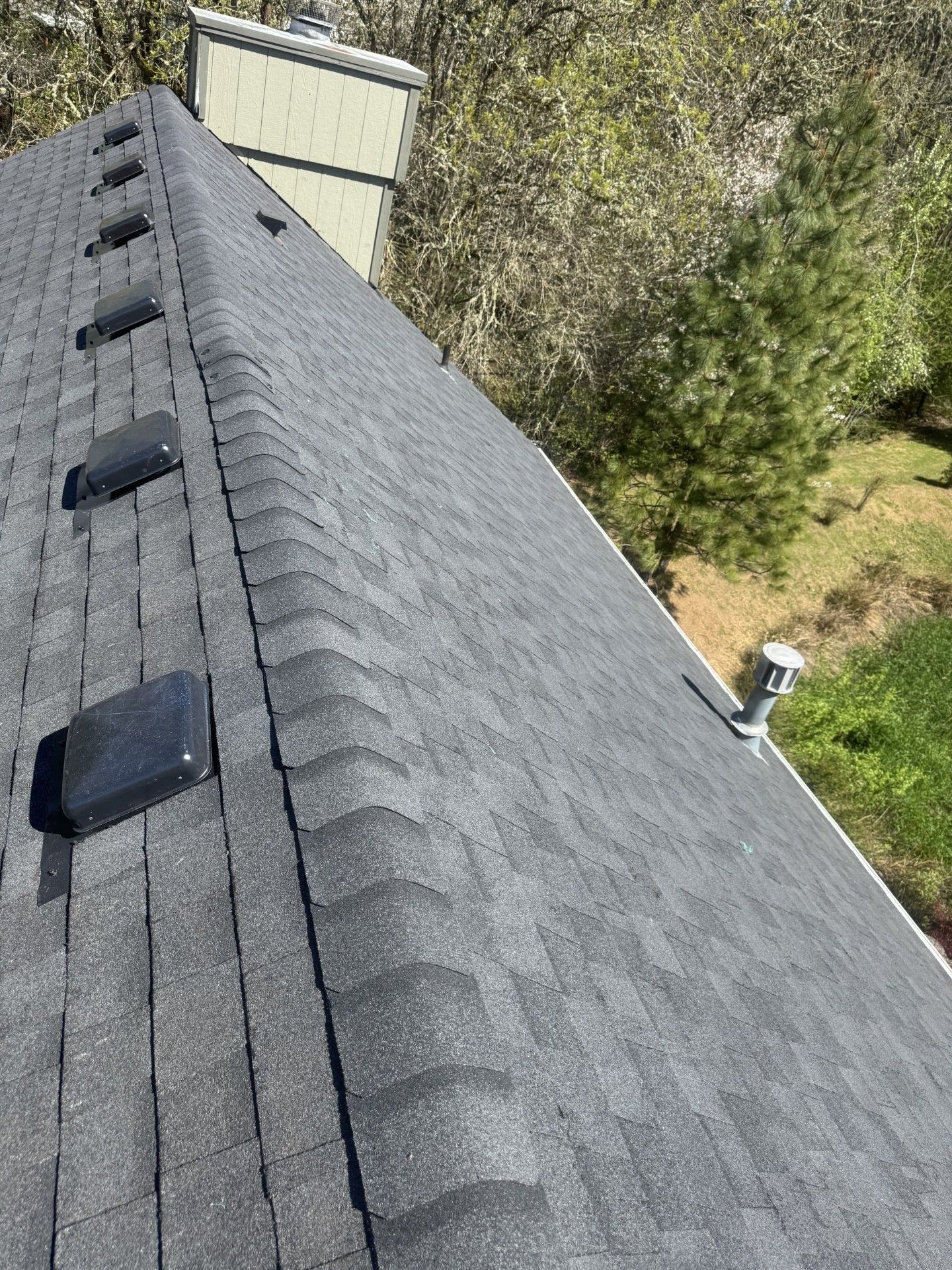 Aerial view of a dark shingled roof with vent pipes and a yard beside it