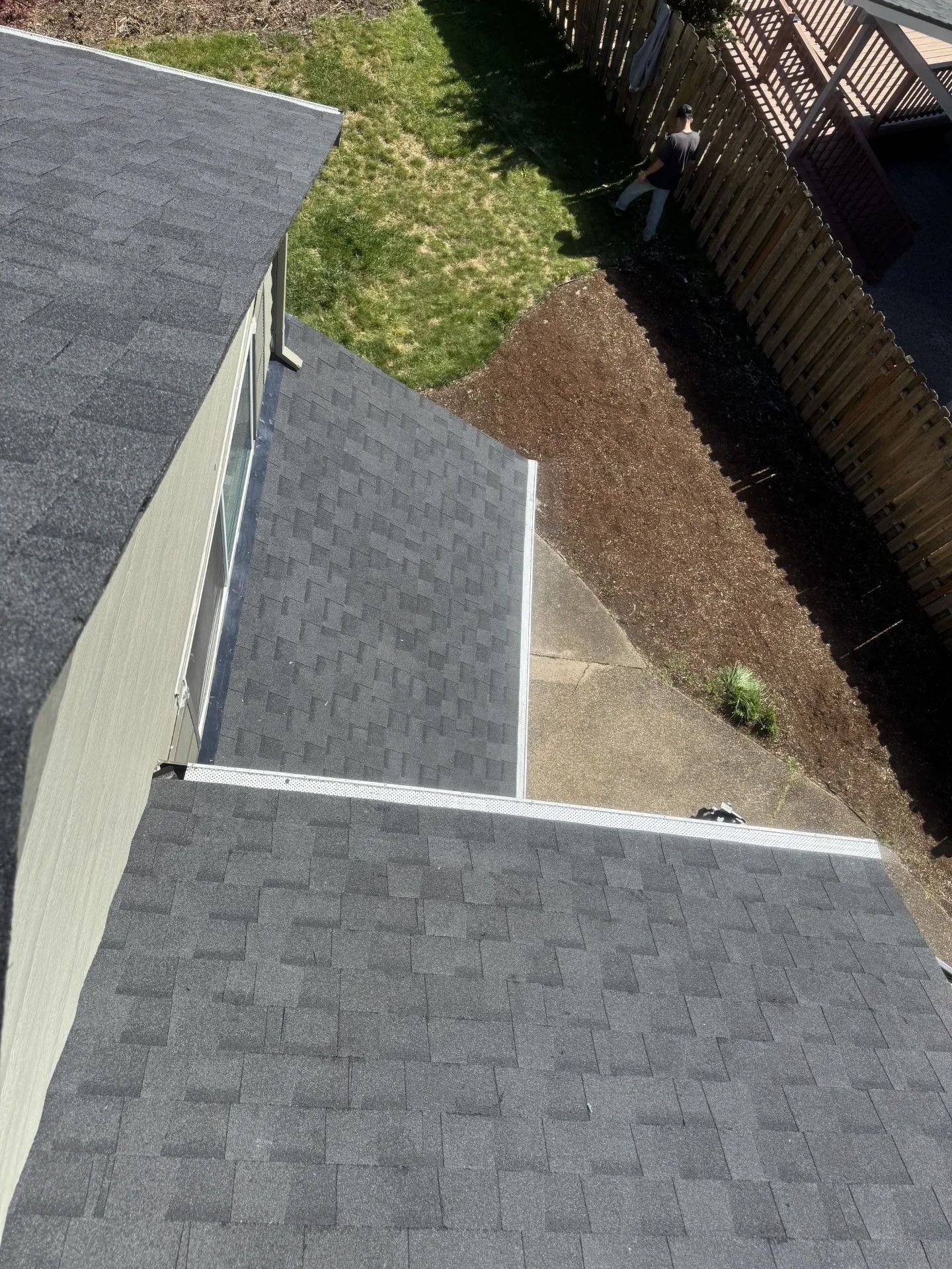 Roof shingles and siding beside a grassy yard and fence, viewed from above