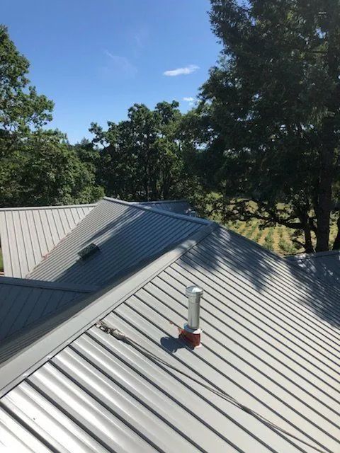 Metal rooftop with a vent pipe and surrounding trees under a blue sky