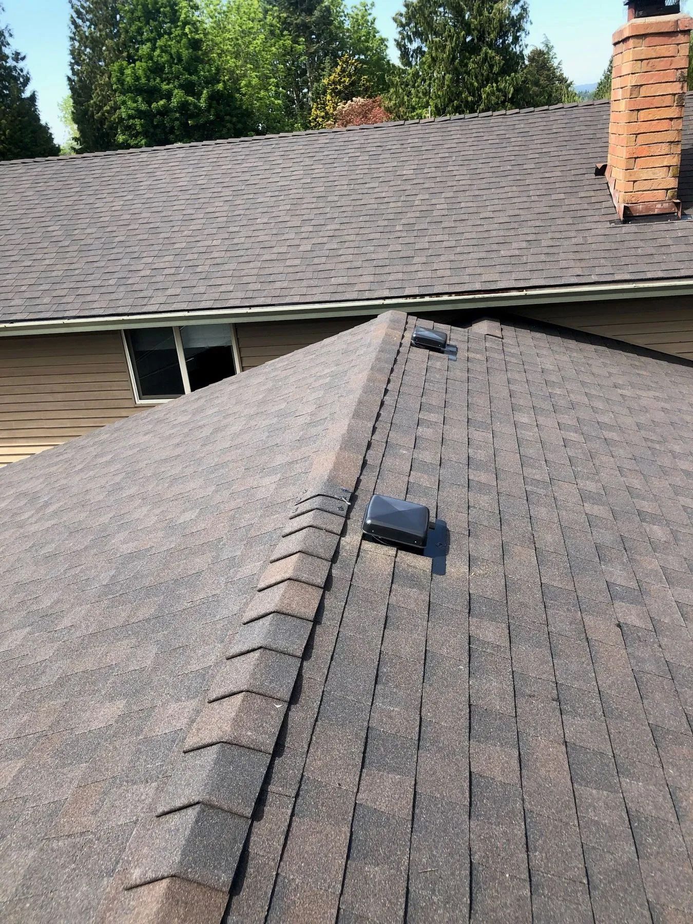 Roof with gray shingles and a central ridge vent on a house near a brick chimney and trees