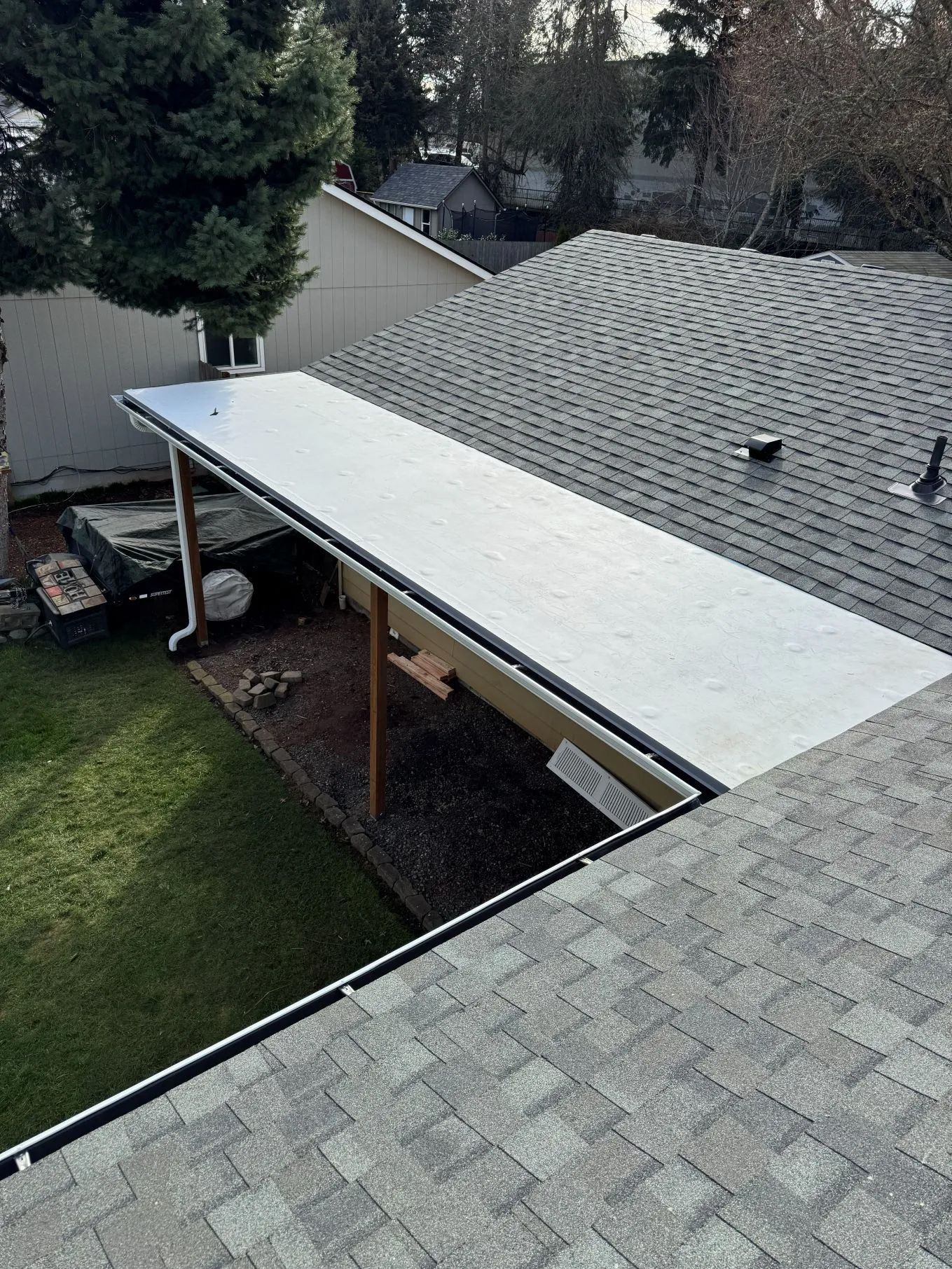 White patio cover attached to a house with gray shingle roof and backyard lawn below