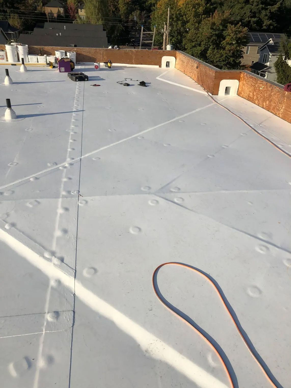 Flat white rooftop with safety lines, vents, and a hose near the brick parapet on a sunny day